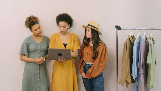 three women standing and looking at a laptop with clothes hanging to the right